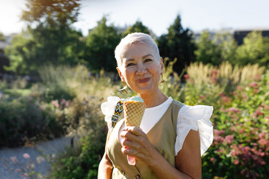 Gorgeous happy senior elegant woman with short haircut eating delicious tasty pistachio ice cream in waffle cone, admiring picturesque views in city park on warm summer evening, enjoying stroll