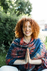 Happy smiling positive redhead millennial girl with curly hair sitting on grass outdoor in city park meditating and practicing yoga, wearing colorful stole on shoulders on chilly early morning air - 785198143