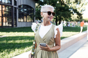 Mature stylish female business couch going to work and walking on her way to office on summer morning, wearing trendy outfit, carrying stylish bag and holding smartphone, looking aside - 785198193