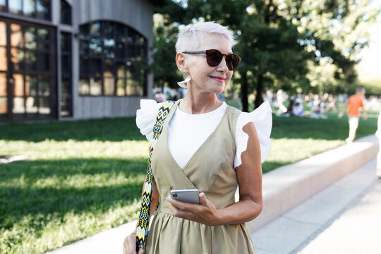 Mature stylish female business couch going to work and walking on her way to office on summer morning, wearing trendy outfit, carrying stylish bag and holding smartphone, looking aside
