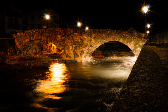 A bridge with a river running underneath it. The bridge is lit up at night. The lights reflect off the water