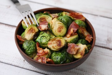 Delicious roasted Brussels sprouts and bacon in bowl on light wooden table, closeup