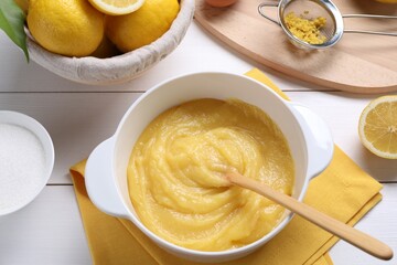 Delicious lemon curd in bowl, ingredients, spoon and sieve on white wooden table, above view