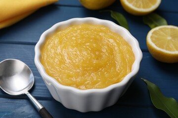 Delicious lemon curd in bowl, fresh citrus fruits and spoon on blue wooden table, closeup