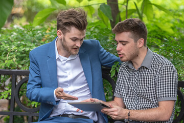 Two businessman or gay friend partner sitting at chair in park while using digital tablet and paper...