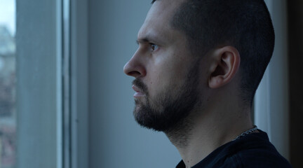 Side view portrait of a thoughtful bearded man looking out the window in bedroom at home.