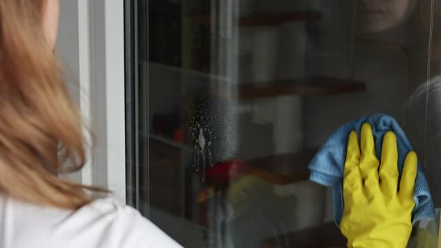 Woman, housework and housekeeping concept - woman in gloves cleaning window with rag and cleanser spray at home