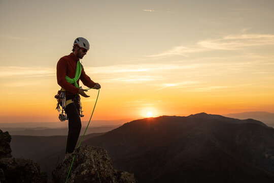 A man is standing on a mountain with a green rope in his hand. The sun is setting in the background, creating a beautiful and serene atmosphere
