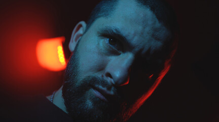 Close-up portrait of a man looking seriously and aggressively directly into the camera, standing against a dark background illuminated by a red light.