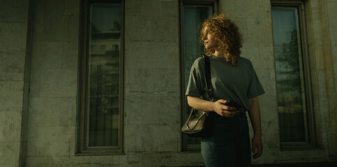 Portrait of a young curly haired woman looking back over her shoulder while holding her smart phone in her hands and standing outside in front of a city
