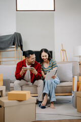 Asian young attractive couple man and woman use tablet and smartphone online shopping furniture decorate house with carton package move in new house.