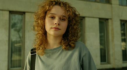 Beautiful young woman with curly hair wearing grey t-shirt looking at camera while walking in the city in warm sunny evening. Portrait of cheerful pretty woman