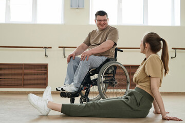 Long shot of mature man in wheelchair and young woman chatting while having break during dance workout
