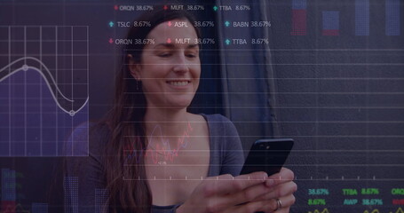 Image of multiple graphs and trading boards, caucasian woman sitting and using smartphone