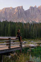 Fototapeta na wymiar White woman in black dress sitting on the shores of an alpine lake surrounded by trees and mountains. A sense of peace and freedom. Lago Di Carezza. Dolomites. Italy