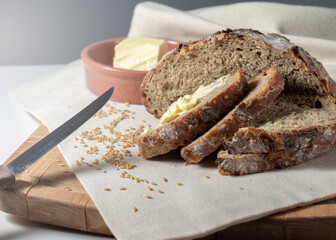 Homemade baking. Round sliced ​​whole grain bread is on a wooden board and butter is in a ceramic plate in blurry focus.