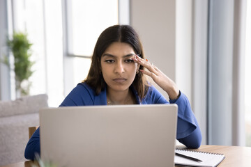 Serious Indian student girl studying at laptop at home, looking at screen, watching learning video lecture, touching head in deep focus. Young freelancer woman thinking on business project