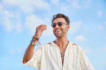 Handsome trendy man wearing a casual shirt, sunglasses, necklace and bracelets looks happy and...