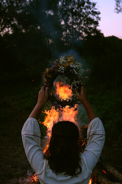 Girl with floral wreath in hands near campfire in forest, dark nature background. rear view. Magic ceremonial, witchcraft. witch wiccan ritual for Beltane, Midsummer, Litha.