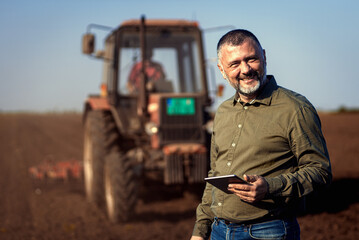 Portrait of satisfied mature farmer standing in field with tablet supervises the cultivation of...
