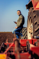 Farmer using tablet while resting after cultivation of soil leaning on tractor tire.