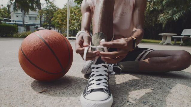 Tilt down shot of African American basketball player sitting on court and tying shoelaces while preparing for game outdoors
