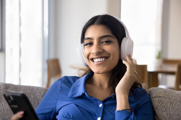 Happy beautiful 20s Indian student girl listening to music from big headphones, holding mobile phone, enjoying online playlist, Internet media musical service, looking at camera with toothy smile