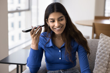 Cheerful young Indian woman holding smartphone at ear, listening to voice message on telephone, smiling, laughing, using media service, internet technology for communication