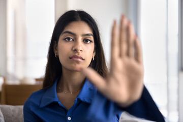 Serious determined young Indian woman showing stop symbol, open palm at camera, making halt, refuse, danger, rejection, restriction hand gesture. Close up of blurred arm with female model
