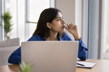 Serious thoughtful Indian freelance woman thinking on work project at laptop, looking at window away, biting thumb in deep thoughts, making decision, pondering on business plan