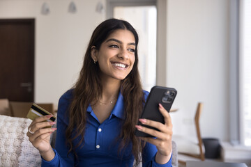Cheerful dreamy young Indian consumer woman enjoying online shopping on mobile phone, paying by credit card, spending money for purchases on Internet, looking away, thinking, smiling