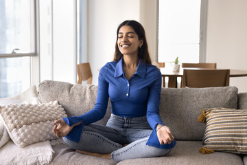 Happy peaceful young Indian woman practicing meditation, breathing at home, meditating on couch, keeping fingers in jnana mudra, getting energy, harmony, training concentration