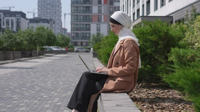 A young woman dressed in a hijab sits on a bench, works on a laptop and waves to a friend. Slow motion. 