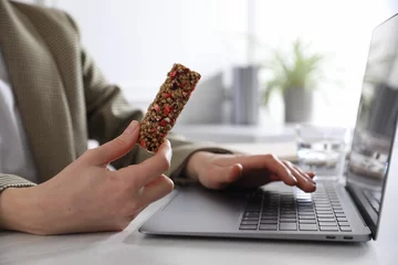 Tuinposter Woman holding tasty granola bar working with laptop at light table in office, closeup © New Africa