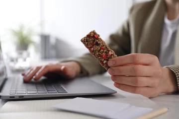 Tuinposter Woman holding tasty granola bar working with laptop at light table in office, closeup © New Africa