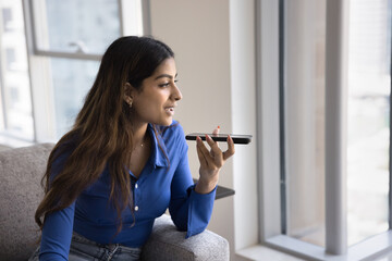 Positive beautiful young adult Indian girl recording voice message on telephone, holding smartphone at mouth, talking on speaker on cell, activating smart home application, looking away