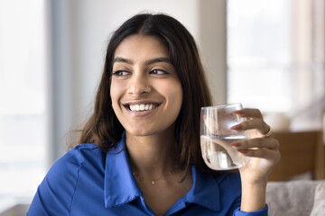 Cheerful beautiful young Indian woman promoting healthy aqua balance, holding glass of cold fresh clean water, looking away with toothy smile. 20s girl caring for healthy lifestyle, skincare