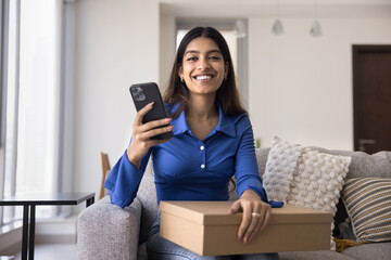 Happy beautiful young Indian shopper girl holding mobile phone and cardboard box with purchase from online store, giving positive feedback on Internet to delivery service, looking at camera, smiling
