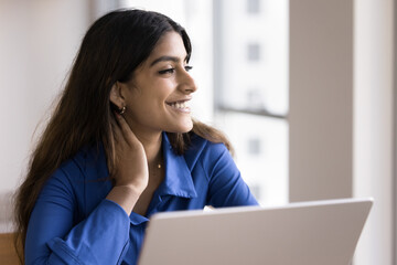 Happy Indian freelance worker girl sitting at laptop, touching neck in deep positive thoughts, looking away, smiling, dreaming, thinking on Internet communication technology