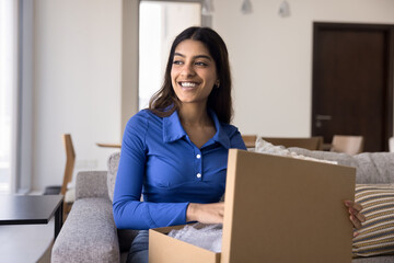 Cheerful young Indian customer girl unpacking parcel at home, holding open cardboard box, getting purchase from online store, looking away with toothy smile, laughing, thinking