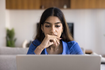Thoughtful young Indian professional girl sitting at workplace table with laptop, working on online business project at home, touching chin, looking at screen in deep focus, using job application