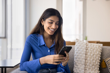 Happy young adult Indian girl using online application on smartphone, sitting on comfortable couch at home, typing message on online chat, enjoying Internet communication, browsing social media