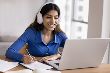 Happy Indian girl in wireless headphones watching learning webinar, online lecture on laptop, speaking at educational conference, writing notes at table, studying on Internet from home