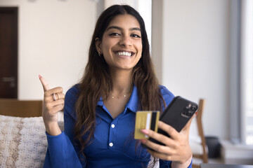 Happy cheerful young Indian customer girl satisfied with bank app holding credit card and smartphone, making like thumb up gesture, looking at camera, smiling, promoting online financial service