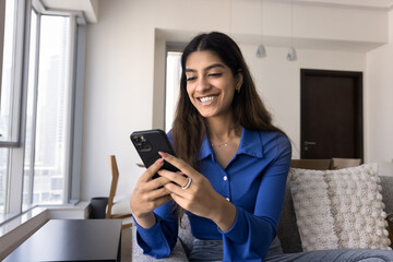 Positive excited young Indian woman typing on mobile phone, using smartphone application, online media v service for entertainment, enjoying internet communication, sitting on couch