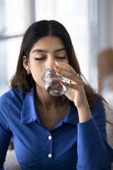 Calm thirsty young 20s Indian girl enjoying cold beverage, drinking fresh clear water with closed eyes, holding glass, promoting healthy lifestyle, diet, detox, caring for aqua balance. Vertical shot