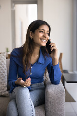 Happy joyful beautiful Indian woman enjoying mobile phone call, communication, conversation, speaking on telephone at home, sitting on couch, looking away with toothy smile. Vertical shot