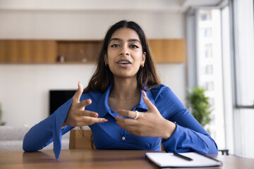 Positive attractive young Indian influencer girl speaking at camera, sitting at work table, moving hands, talking on video conference call about business project, broadcasting on Internet
