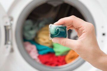 Young adult woman hand holding blue liquid capsule for colorful clothes washing on washing machine background. Closeup. Front view.