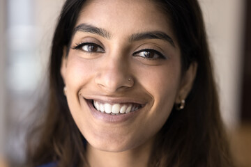Happy beautiful young 20s Indian girl face close up. Cheerful female model, woman with healthy white teeth, smooth clean skin and makeup looking at camera for cropped portrait - obrazy, fototapety, plakaty